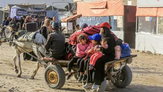 three young girls and a women sit on the back of a trailer pulled by a horse. all their belongings are also on the trailer. There is a man sitting at the front guiding the horse.