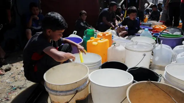 Palestinian children fill water containers at a tent camp, boys sat near lots of buckets.