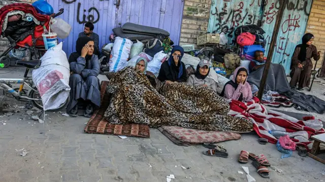 five women sit under blankets resting on their belongings that are leaning against a wall outside. Some distance from them, a two separate men also with their belongings