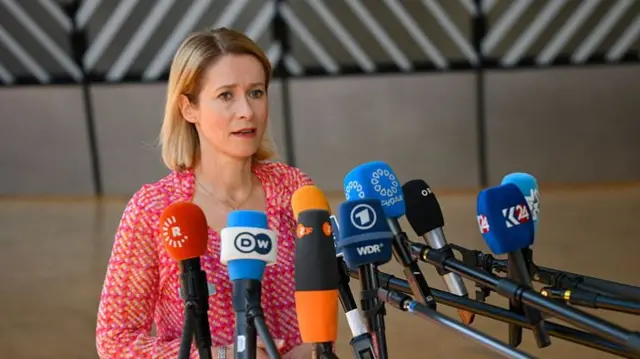 Kaja Kallas standing talking outside EU HQ. There are multiple outlet mics in front of her. She is wearing a pink, white and yellow top.