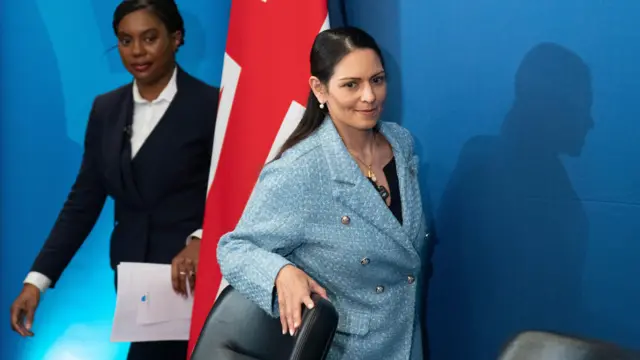 Patel walking towards a chair, with Kemi Badenoch standing behind her