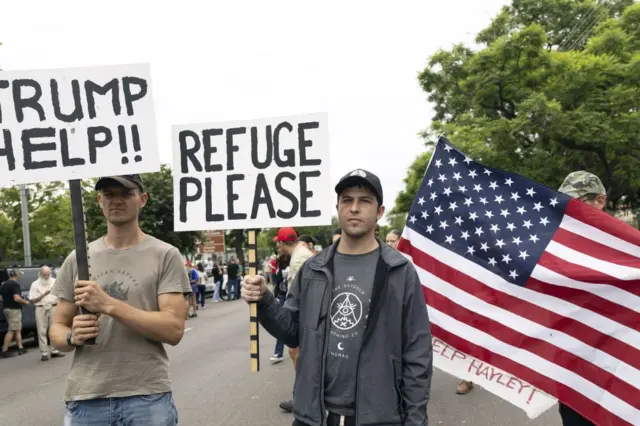 Two protesters, one holding a sign which says: "Refuge please" and another which says "Trump help"