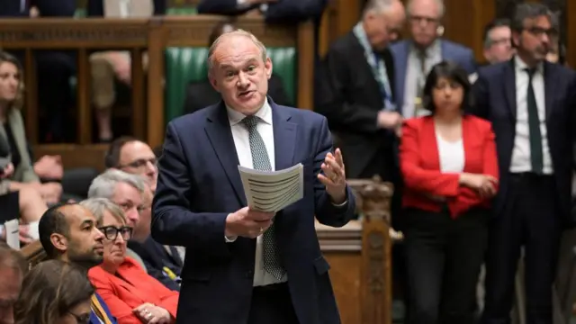 Ed Davey gestures while talking and holding some papers in the House of Commons.