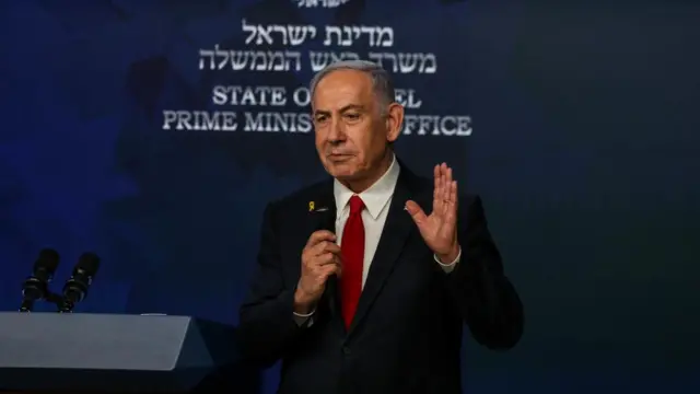PM Netanyahu speaks in front of a sign saying PM's Office, wearing a suit and holding a mic.