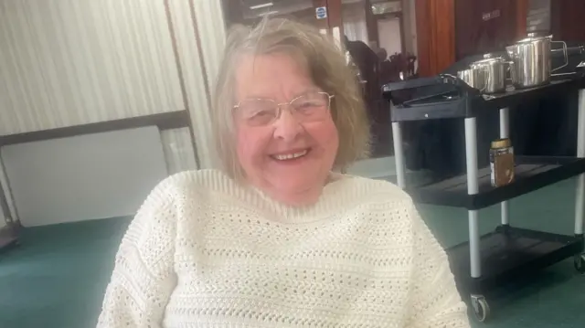 An elderly woman smiles at the camera while seated.