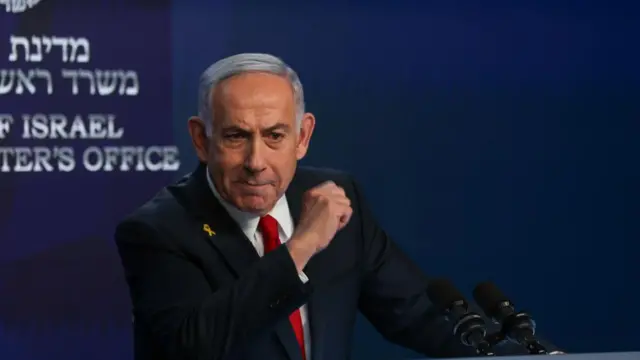 Netanyahu clenches his fist and speaks behind a podium, wearing a suit and red tie.