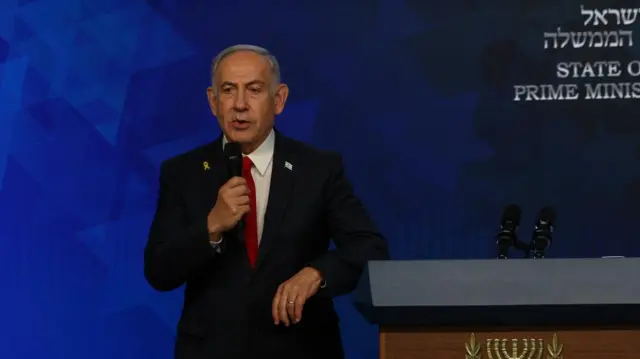 Netanyahu speaks holding a microphone behind a podium wearing a suit.