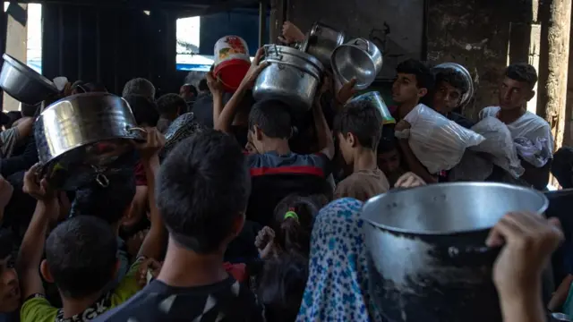 Hundreds of displaced Palestinians gather outside the Sokar Charity Kitchen in Gaza City to receive limited food rations. Many children hold steel pans in the air and are crowded in a room.