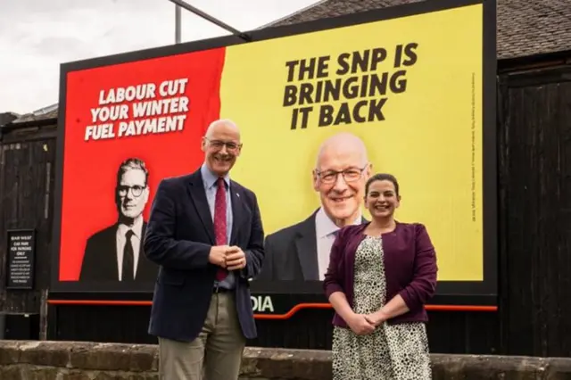 A photo of John Swinney smiling next to a billboard with a caption which reads: "A reminder to everyone in Scotland that Labour cut the winter fuel payment for pensioners and the SNP brought it back."
