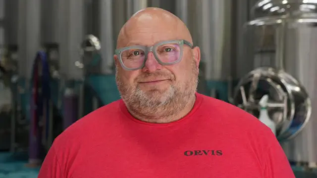 James Cuthbertson wearing a red tshirt and spectacles looks directly at the camera with brewery equipment behind him in the background