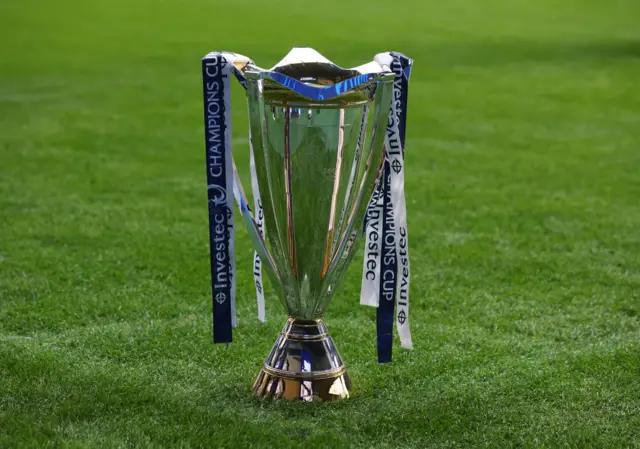 The European Champions Cup trophy on a grass pitch. The trophy is decorated with blue and white ribbons which carry the name of tournament sponsor Investec