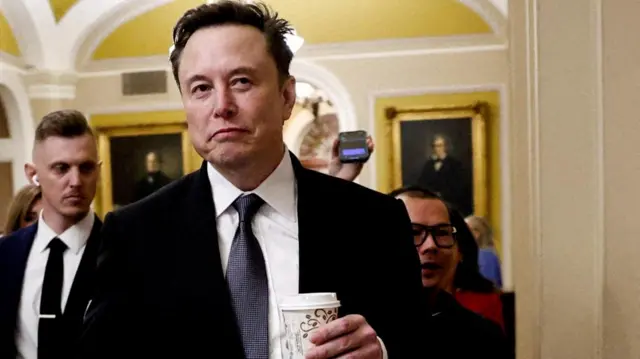 Musk, wearing a suit, walks through the halls of the White House carrying a disposable coffee cup
