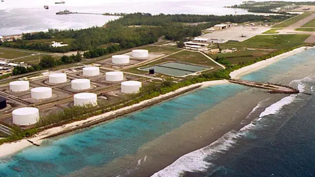 An overhead image shows fuel tanks on a strip of land surrounded by the sea