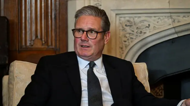 Keir Starmer wears a black suit and is sitting, wearing glasses.