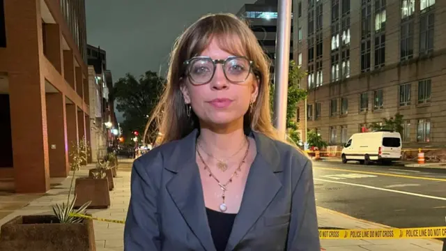 A woman in a suit and glasses looks into the camera. She wears two necklaces.