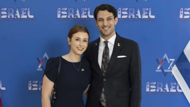The young couple pose together, each with one arm around the other, wearing smart dress at what appears to be a formal occasion - they are standing in front of a board that says "embassy of Israel to the US" on it, with an Israeli and US flag on either side