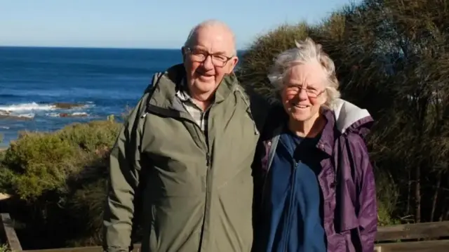 Don Patterson, who is bald and wears glasses and a green jacket, stands with his wife Gail, who has grey hair and glasses and is wearing a blue fleece and purple rain jacket. They are standing in front of the sea, as seen from what looks like a coastal walk.