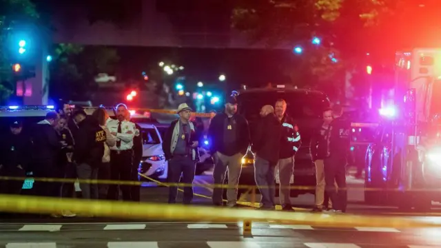 Police officers stand at the scene, with the lights of emergency vehicles lighting the scene red and blue