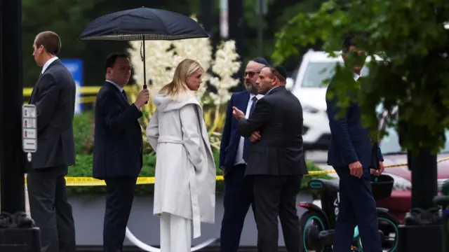 US Attorney General Pam Bondi stands at the scene with yellow police tape behind her. She is peaking with two men