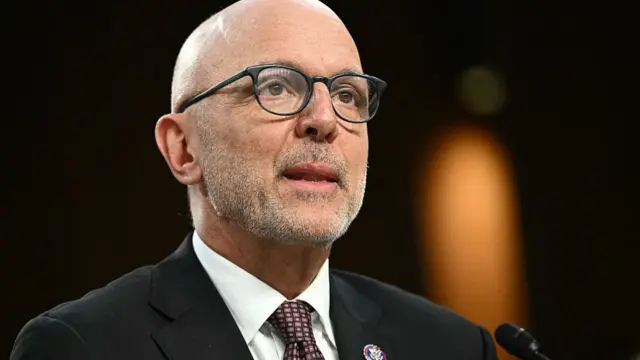 Close up of Ted Deutch's face, speaking indoors, wearing a suit and glasses with grey stubble