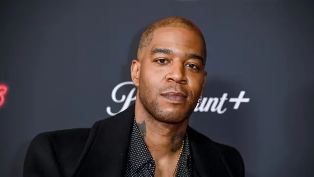 Scott "Kid Cudi" Mescudi is looking at the camera. He is wearing a black jacket over a black polka dotted shirt and a chain.