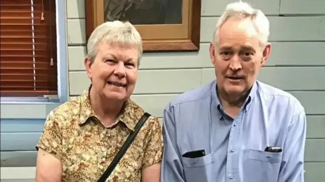 Heather Wilkinson, wearing a yellow floral shirt, smiles at the camera next to her husband Ian, who is wearing a blue shirt with things in the pockets. They both have grey hair.