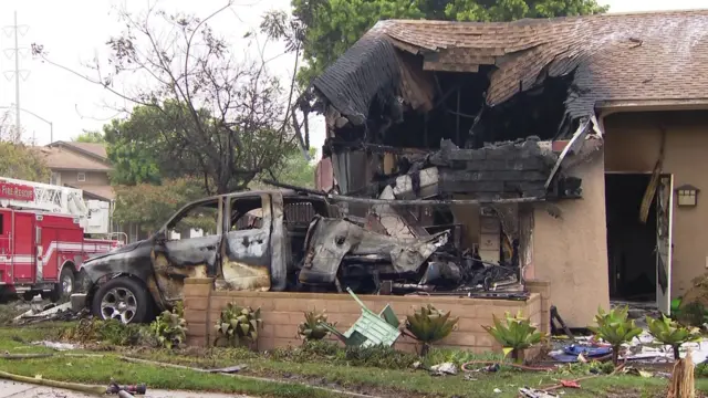 The roof of a house caved in with a burnt out car next to it, and a fire engine