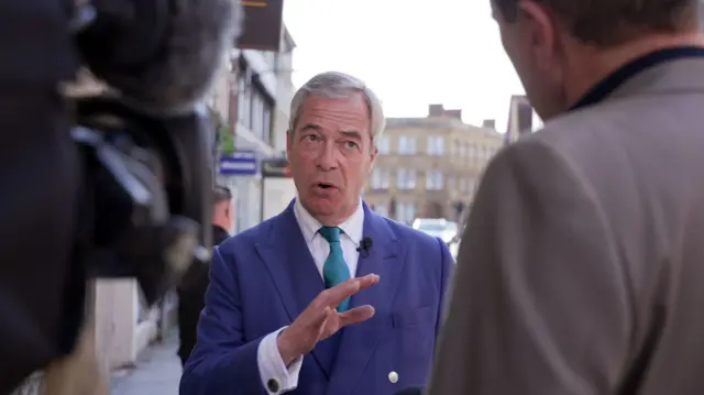 An image of Nigel Farage speaking to the media. He faces towards the camera while the outline of a camera and a reporter can be seen at the edges of the frame
