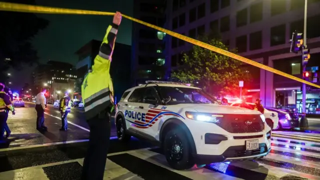 A policeman holds up police tape at the scene to allow a police car to drive underneath. Other officers and police vehicles can be seen behind