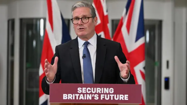Keir Starmer speaks, gesturing his hands. three union flags can be seen behind him. he is standing at a purple lectern that reads 'securing britain's future'. he is wearing a black suit and black glasses.
