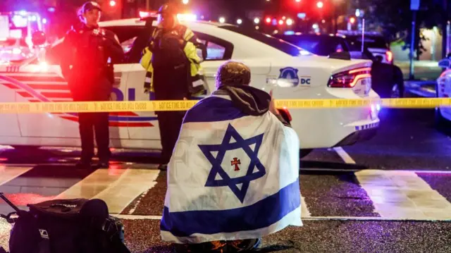 A person draped in an Israeli flag which had a small red cross which says 'Jesus' on it. They kneel at the scene with a police car, officers and police tape in front of them