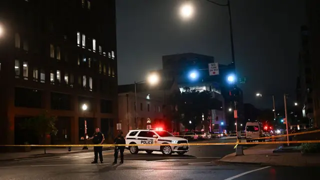 Two police officers stand at the scene, with their vehicle behind them and the street marked off by police tape. Unlike the other pictures, the street is very empty