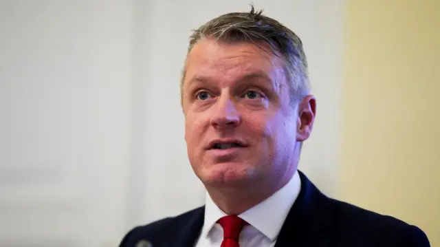 Pollard speaks as he looks forward off to the side of the camera. You can see his shoulders and above, he wears a suit, white shirt and red tie. The background is beige and blurry
