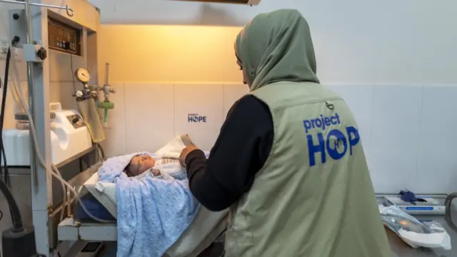 Project Hope worker, standing next to a baby sleeping in a hospital cot