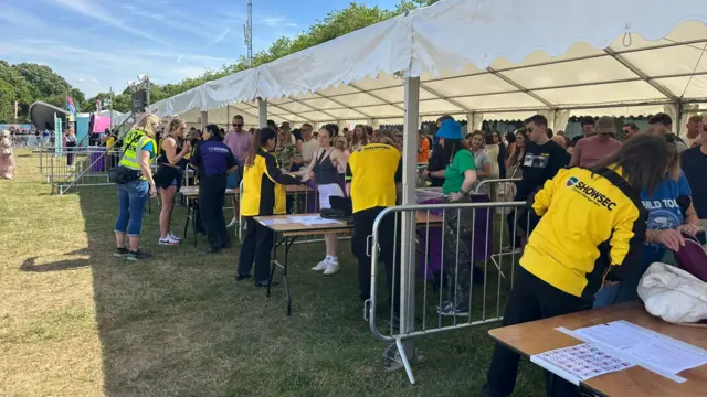 People stand in a queue under a white canopy while security officials carries out checks.