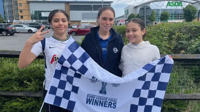 Soraia (L), Charlene (C) and Shayla (R) are seen standing together holding a commemorative "Euro League Winners" flag. Soraia is making a V for victory sign