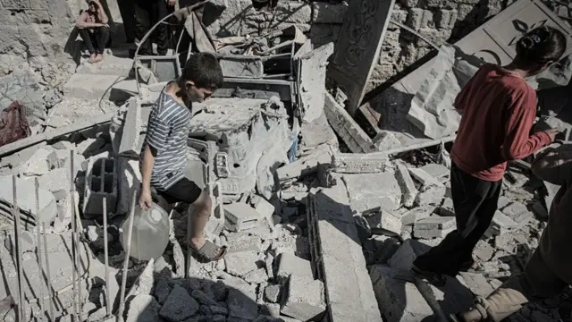 A young boy carrying a large bottle of water covered in dust walks over the debris of a home, a young girl in a red top walking in front of him