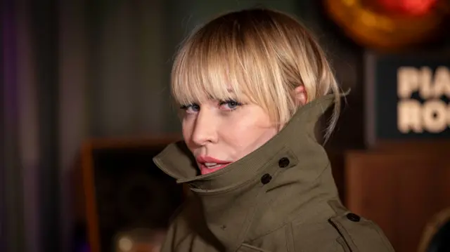 Natasha Bedingfield looks a the camera. She is wearing a khaki coat. She has blonde hair and blue eyes.