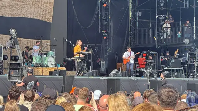 People at Big Weekend watching The Wombats on stage, who are playing a piano, guitar, and drums.