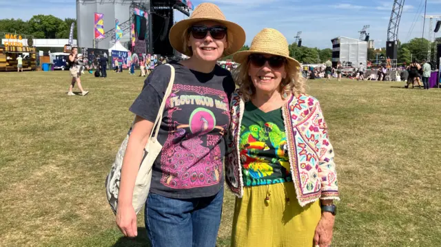 Hannah poses for the camera with Emma at Big Weekend. They are both wearing straw hats and colourful graphic t-shirts.