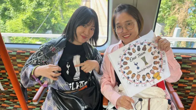 Two Korean women displaying Tottenham merchandise sitting on a train, The one on the left is wearing a dark Tottenham Hotspurs jersey under a flannel shirt and pointing at their team logo with her left pointer finger