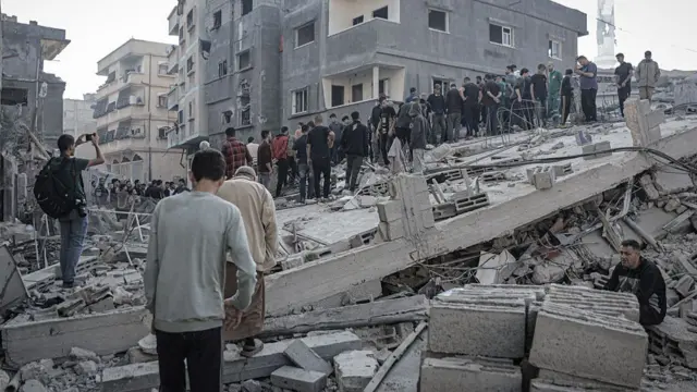 A large group of men in joggers stand on the rubbles of a house in Gaza, debris filling the foreground and a group of damaged homes in the background
