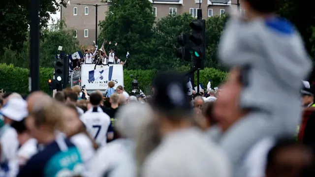 Crowds in the foreground are seen watching a cheering Spurs team on the open-top bus