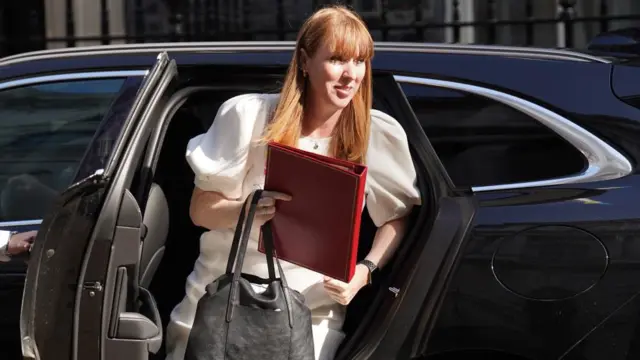 Rayner exiting a black car on Downing Street, holding a red folder and a handbag. Wearing a white dress and smiling.