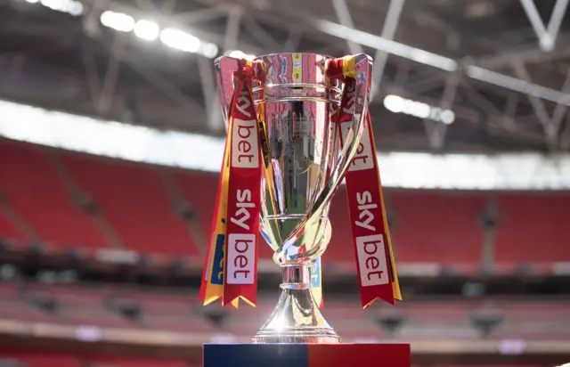 The Championship play-off final trophy. It is silver and there are red, white and yellow ribbons tied around the handles of the trophy that read 'Skybet'. It is standing against the backdrop of Wembley Stadium.