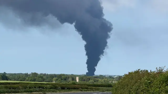 A plume of black smoke rises from the ground against a blue sky. A road surface is visible in the foreground