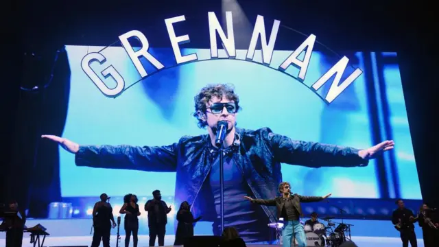 Tom Grennan extends his arms on stage. There's a screen that shows him in the background.