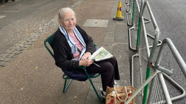 Kathleen Sleap is pictured in a camping chair with a newspaper in hand in front of barriers along the parade route