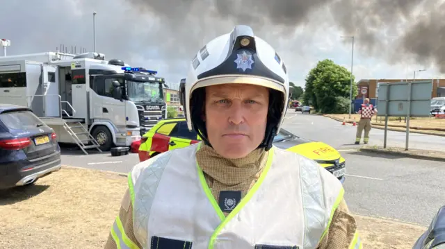 Mick Berry, in firefighter uniform and a helmet stares straight to the camera. Smoke from the fire is partially visible over his shoulder
