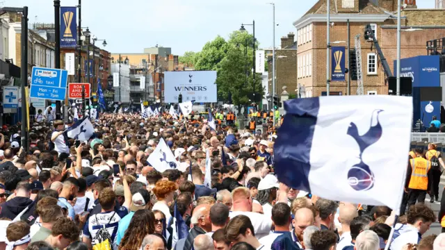 A dense crowd of thousands Spurs fans are seen. In the distance is a screen that reads: winners. Blue and white flags with the team's crest are seen throughout